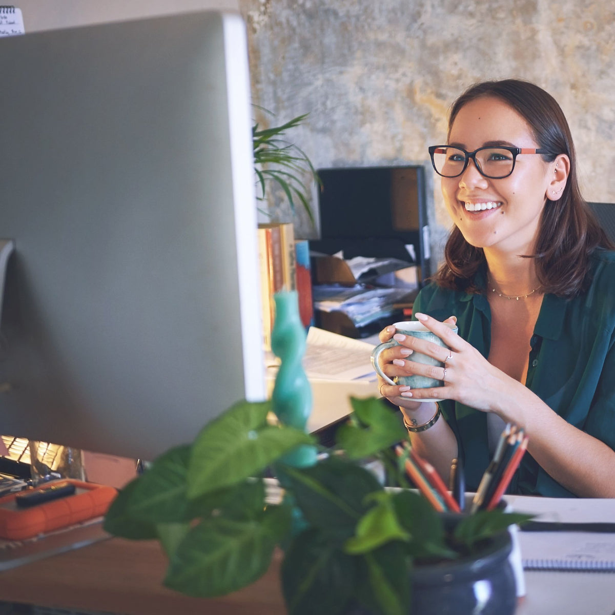 A woman with glasses and a green shirt smiles while holding a mug at her desk in a modern office setting, with a large computer monitor in the foreground.